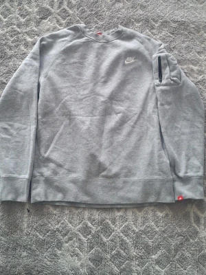 Grå sweatshirt från Nike - En stilren grå sweatshirt från Nike med klassisk logga på bröstet. Tröjan har rund halsringning, långa ärmar och en praktisk ficka med dragkedja på ena ärmen. Perfekt för en avslappnad och sportig look.