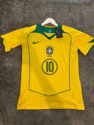  Ronaldinho - Gul Brasilien fotbollströja från Nike med gröna detaljer, nummer 10 och Ronaldinho på ryggen. Klassisk landslagströja med broderad logga och meshpaneler för ventilation. Perfekt för dig som älskar fotboll och vill bära en ikonisk matchtröja.