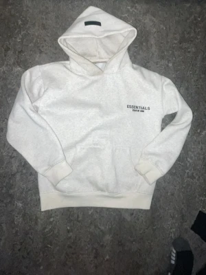 Vit Essentials Fear of God hoodie - Snygg vit hoodie från Fear of God Essentials med svart logga tryckt på bröstet och ryggen. Klassisk passform med huva och känguruficka framtill. Perfekt för en clean och stilren streetwear-look. använt typ åtta gånger
