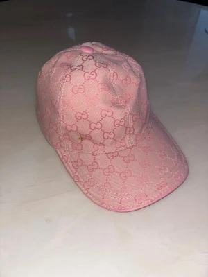 Gucci keps - Snygg rosa keps från Gucci med klassiskt GG-monogram över hela kepsen. Kepsen har böjd skärm och är tillverkad i ett tygmaterial med ton-i-ton detaljer. Perfekt accessoar för att lyfta din outfit.