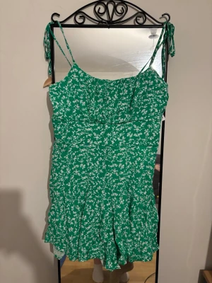 Grön blommig kortklänning med knyt - Superfin grön jumpsuit med vita småblommor och smala axelband som knyts på axlarna. Lätt och luftig modell med markerad midja och korta ben. Perfekt för varma dagar.