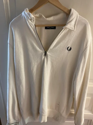 Vit halfzip hoodie från Fred Perry - Snygg vit hoodie från Fred Perry med halv dragkedja och broderad logga på bröstet. Tröjan har långa ärmar, ribbade muddar och är gjord i mjukt material. Perfekt för en clean och stilren look.