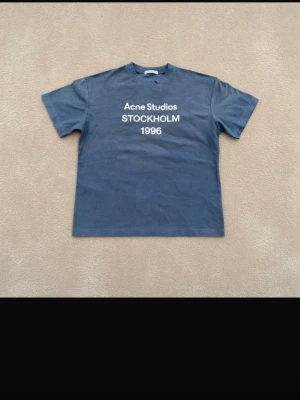 Blå Acne Studios t-shirt - Snygg blå t-shirt från Acne Studios med vit text 'STOCKHOLM 1996' på bröstet. Klassisk passform och rund halsringning. Perfekt för dig som gillar stilrena och trendiga plagg med streetkänsla. äkta !!!