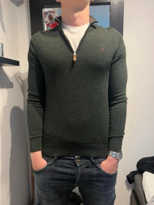 Mörkgrön halvzip-tröja från Morris - Snygg mörkgrön stickad tröja från Morris med halv dragkedja och liten broderad logga på bröstet. Tröjan har ribbade muddar och är figurnära i modellen. Perfekt att bära över en t-shirt för en stilren look.