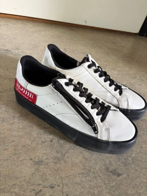 Vita sneakers med svart sula och dragkedja - Snygga vita sneakers med svart sula, svarta skosnören och dragkedja på sidan. Skorna har en röd detalj med vitt mönster vid hälen och svart innerfoder. Perfekta för dig som gillar en unik och sportig stil.