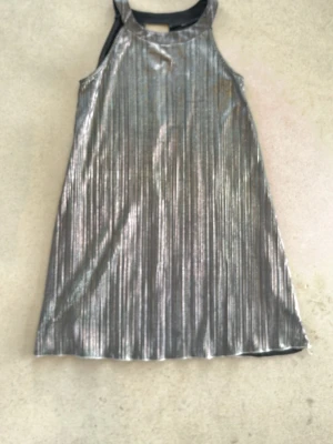 Glittrig silverklänning i A-linje - Säljer en glittrig silverfärgad klänning i A-linjeform med ribbade vertikala ränder. Klänningen är ärmlös och har en hög halsringning. Perfekt för dig som vill sticka ut med metallic vibes.