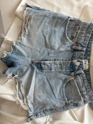Ljusa denimshorts från Zara - Säljer ett par ljusa denimshorts från Zara med råa, fransiga kanter och klassisk femficksdesign. Shortsen har hög midja och är gjorda i bomull. Perfekta för sommarens alla äventyr och har en avslappnad, loose passform.