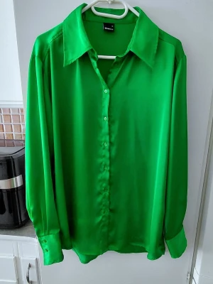 Grön satinliknande skjorta - Säljer en grön skjorta i satinliknande material med klassisk krage och knappar framtill. Skjortan har långa ärmar med breda manschetter och en loose passform som ger en avslappnad men ändå stilren look. Storlek M. Fint skick. 