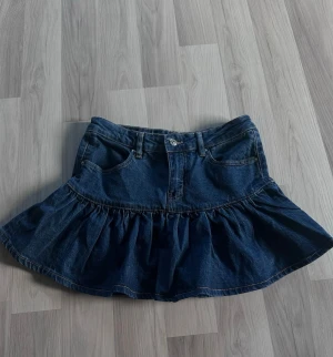 Mörkblå volangkjol i denim - Snygg mörkblå jeanskjol med volang och klassiska fickor fram och bak. Kjolen har en markerad söm och knappstängning framtill. Perfekt för dig som gillar en trendig och avslappnad stil.