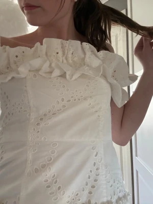 Vit offshoulder blus med volang - Supersöt vit offshoulder blus med broderade detaljer och volang runt hela överdelen. Blusen är ärmlös och har ett romantiskt, somrigt uttryck med fina hålmönster. Perfekt för dig som vill ha en feminin och trendig topp.