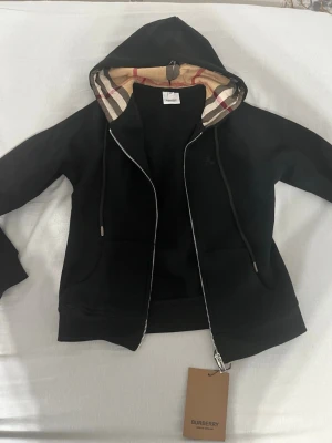 Svart hoodie från Burberry med dragkedja - Svart hoodie från Burberry med klassiskt rutigt foder i huvan, dragkedja framtill och två fickor. Stora snören i huvan med metalländar ger en snygg detalj. Perfekt för en stilren och lyxig streetlook.