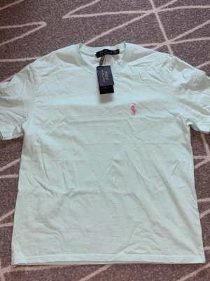 Vit t-shirt från Polo Ralph Lauren - Fräsch vit t-shirt från Polo Ralph Lauren med klassisk passform och korta ärmar. T-shirten har en liten rosa broderad logga på bröstet och är tillverkad i mjuk bomull. Perfekt till jeans eller shorts.