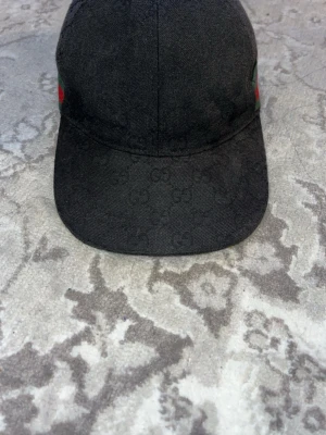 Svart keps från Gucci - Snygg svart keps från Gucci med diskret GG-logomönster över hela kepsen. På sidan finns det klassiska gröna och röda ränderna. Kepsen har böjd skärm och är tillverkad i tygmaterial. Perfekt för dig som gillar exklusiva accessoarer. Dustbag kan slänga med om så önskas!