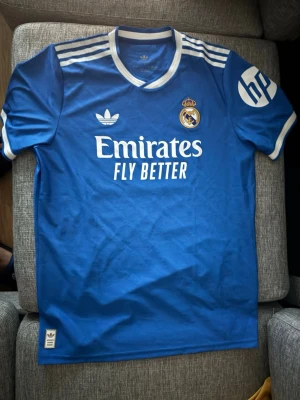  Real Madrid third jersey 2025/2026 (Mbappe 10) authentic  - Snygg blå Real Madrid matchtröja från Adidas med vita detaljer och klubbmärke på bröstet. Tröjan har vita ränder på axlarna, HP-logga på ärmen och Emirates Fly Better-tryck på framsidan. Perfekt för fotbollsfans som vill sticka ut på läktaren eller planen.