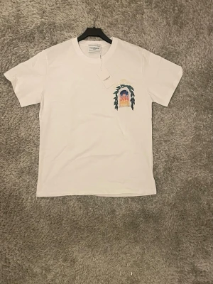 Vit t-shirt från Casablanca - Vit t-shirt från Casablanca med färgglad tryckt grafik på bröstet och ryggen som föreställer en portal och texten 'Avenida Casablanca'. Klassisk passform och rund halsringning. Perfekt för dig som gillar stilrena men unika plagg.