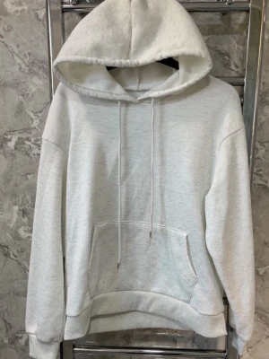 Ljusgrå basic hoodie med huva - En stilren ljusgrå hoodie med huva och dragsnören. Hoodien har en klassisk känguruficka framtill och ribbade muddar vid ärmar och nederkant. Perfekt för en avslappnad och enkel look. Materialet ser ut att vara mjukt och bekvämt, troligen bomullsblandning.