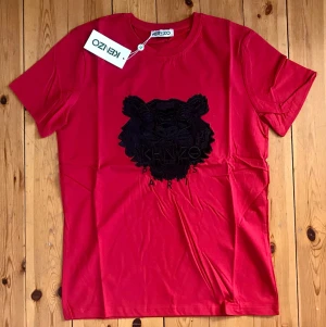 Röd Kenzo t-shirt med tigertryck - Säljer en röd t-shirt från Kenzo med det ikoniska svarta tigerhuvudet broderat på bröstet. T-shirten har rund halsringning och korta ärmar. Tillverkad i mjuk bomull som känns skön mot huden. Perfekt för dig som vill sticka ut med en klassisk streetwear-look.