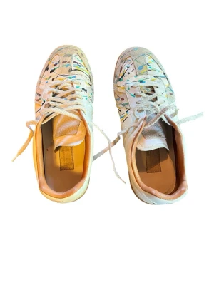 Vita sneakers med färgstänk - Unika vita sneakers med färgstänk i blått, gult och svart. Skorna har lågt skaft, vita snören och beige innersula. Ovandelen är i läder och mocka, vilket ger en schysst mix av material och textur. Perfekta för dig som vill sticka ut med din stil.