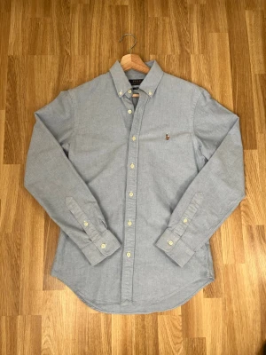 Blå skjorta från Polo Ralph Lauren - Klassisk ljusblå skjorta från Polo Ralph Lauren med button-down krage och broderad logga på bröstet. Slim fit-modell i bomull med långa ärmar och vita knappar. Perfekt för en clean och stilren look.