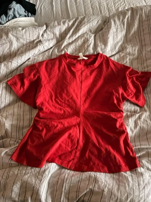 Röd kortärmad topp med sömdetalj - En röd topp med korta ärmar och rund hals. Toppen har en markerad söm som går vertikalt både fram och bak, vilket ger en cool och modern look. Materialet känns som bomull och passformen är normal. Perfekt för dig som gillar färg och enkel stil.