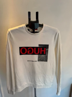 Vit sweatshirt från Hugo Boss storlek S - Vit sweatshirt med rund hals och lång ärm från Hugo Boss. Stor röd och svart logga framtill med texten 'HUGO'. Mjuk bomullskänsla och avslappnad passform. Perfekt för en trendig streetwear-look.