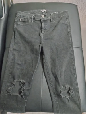 Svarta ripped jeans River Island - Snygga svarta jeans från River Island med slitna hål vid knäna för en trendig look. Klassisk femficksmodell med knappgylf och smal passform. Perfekta för dig som gillar en edgy stil.