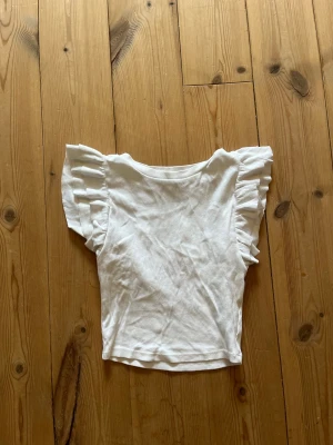 Vit topp med volangärmar - Söt vit topp med breda volangärmar och rund halsringning. Tillverkad i ett ribbat, mjukt tyg som ger en bekväm känsla. Perfekt för dig som gillar en feminin och trendig stil.