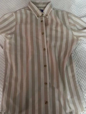 Randig skjorta från Jack & Jones - Säljer en snygg skjorta från Jack & Jones med beige och vita vertikala ränder. Skjortan har klassisk krage, knappar framtill och korta ärmar. Perfekt för en stilren look och tillverkad i mjuk bomull. MARKERA= 176 cm mycket rba skick