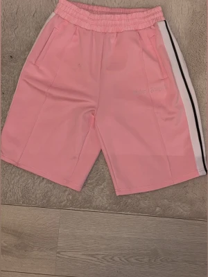 Palm Angels shorts  - Palm Angels shorts i ljusrosa med vit och svart sidoremsa. Elastisk midja och två fickor framtill. Broderad logga på ena benet. Perfekta för en avslappnad och trendig look. Liten fläck på shortsen men går säkert bort om man tvättar. Shortsen är lite baggiga Storlek M passar Även L