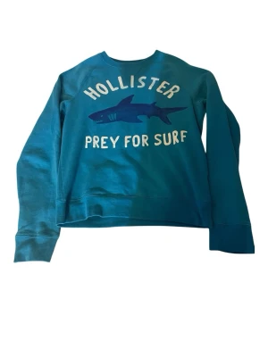 Turkos sweatshirt från Hollister - Cool turkos sweatshirt från Hollister med tryck av en haj och texten 'Prey for Surf' på framsidan. Tröjan har rund halsringning och långa ärmar. Perfekt för en avslappnad och sportig stil.