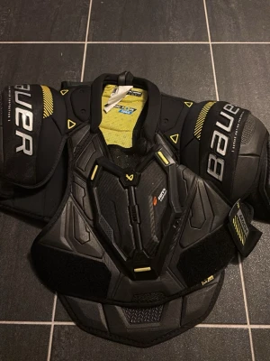 Bauer axelskydd SR Medium mach - Säljer ett par svarta axelskydd från Bauer i storlek SR Medium. Skydden har gula detaljer och är utrustade med Thermo Core Zero-teknologi för extra komfort. Perfekta för hockey eller annan kontaktsport. Snygg design och bra passform.