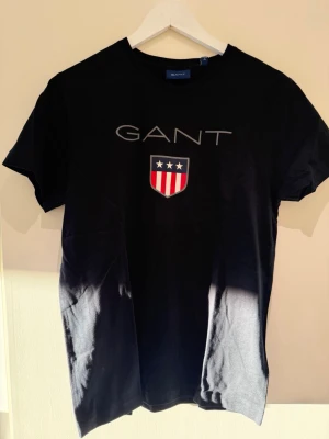 Svart GANT t-shirt med logga - Snygg svart t-shirt från GANT med klassisk logga och sköld med stjärnor och ränder på bröstet. T-shirten har rund hals och korta ärmar, perfekt till jeans eller shorts. Tillverkad i mjuk bomull för skön känsla hela dagen.