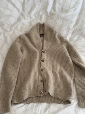 Beige kofta från Massimo Dutti - Säljer en beige ribbstickad kofta från Massimo Dutti med fem knappar framtill och bred krage. Koftan är gjord av italienskt garn och har långa ärmar samt en avslappnad passform. Perfekt för lager på lager och riktigt mysig.