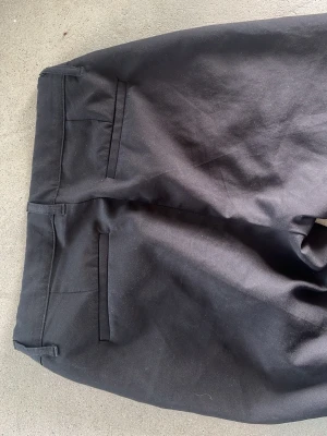 Svarta kostymbyxor från Gant - Snygga svarta kostymbyxor från Gant med klassisk rak passform och två bakfickor. Tillverkade i bomull och elastan för en bekväm känsla och bra stretch. Perfekta för en stilren look och passar till skjorta eller t-shirt.