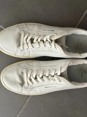 Vita PS Paul Smith sneakers - Stilrena vita sneakers från PS Paul Smith i läder med klassisk låg profil och vita snören. Diskret logga på sidan och hälparti i mörkblått. Perfekta för en clean och enkel look.