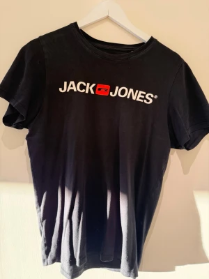 Svart Jack & Jones t-shirt med logga - Snygg svart t-shirt från Jack & Jones med stor vit logga och röd detalj på bröstet. Klassisk rund hals och korta ärmar. Tillverkad i mjukt bomullsmaterial som känns skönt mot huden. Perfekt för en avslappnad och trendig stil.
