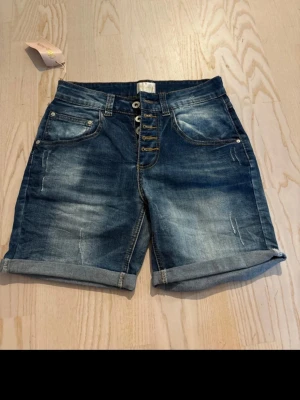 Blå denimshorts med knappar - Snygga blå denimshorts från KappAhl med uppvikta benslut och fem knappar framtill. Klassisk femficksmodell med slitningar och kontrastsömmar. Perfekta för varma dagar och enkel att matcha med olika toppar.