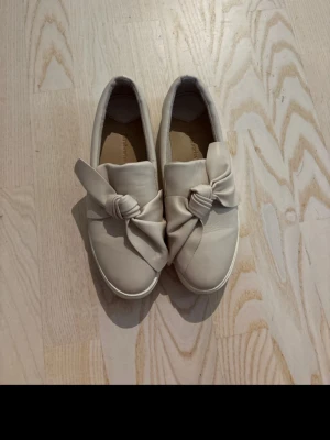 Beige loafers med knutdetalj - Säljer ett par beige loafers med snygg knutdetalj framtill. Skorna har rund tå, vit sula och är tillverkade i ett mjukt material som ser ut som skinn. Perfekta för dig som gillar en stilren men ändå unik look.