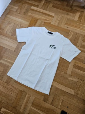 Vit t-shirt från A.P.C. x Lacoste - Snygg vit t-shirt från A.P.C. i samarbete med Lacoste. T-shirten har en klassisk passform, rund hals och korta ärmar. På bröstet finns en grön krokodillogga med texten A.P.C. Tryckt i bomull som känns mjuk och skön mot huden.
