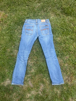 Ljusblå jeans från Nudie Jeans - Säljer ett par ljusblå jeans från Nudie Jeans med klassisk femficksdesign och snygga kontrastsömmar. Jeansen har raka ben och ikoniska broderade bakfickor. Perfekta för en avslappnad och trendig stil. Model Grim Tim