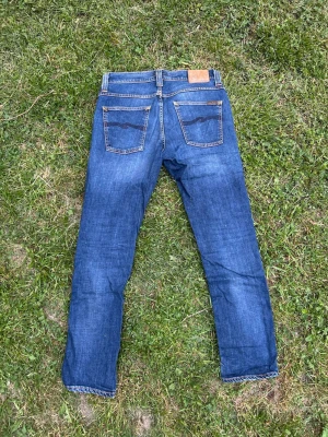 Blå jeans från Nudie Jeans - Säljer ett par klassiska blå jeans från Nudie Jeans med raka ben och fem fickor. Jeansen har snygga kontrastsömmar och en läderpatch bak i midjan. Perfekta till vardags och passar till det mesta. Materialet är slitstarkt denim i bomull. Model Grim Tim 