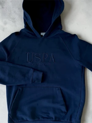 Svart hoodie från US Polo Assn - Snygg svart hoodie från US Polo Assn med stor USPA-logga broderad på bröstet. Klassisk känguruficka framtill och huva. Tillverkad i mjuk bomullsblandning som är skön att ha på sig. Perfekt för en avslappnad och stilren look. Passar både M/S men kan bli lite tajt för M. Fraktar samma dag 📦💨