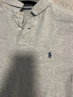 Grå långärmad piké från Ralph Lauren - Klassisk grå långärmad pikétröja från Ralph Lauren med blå broderad logga på bröstet. Tröjan har krage, knäppning med två knappar och är tillverkad i mjuk bomull. Perfekt för en stilren och avslappnad look.