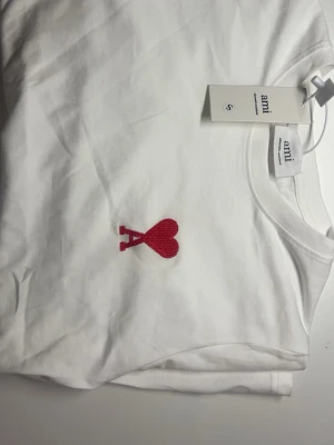 Vit t-shirt från Ami med rött hjärta - Vit t-shirt från Ami med ett broderat rött hjärta och bokstaven A på bröstet. Klassisk rund halsringning och korta ärmar. Tillverkad i mjuk bomull för en skön känsla. Perfekt för en stilren och trendig look.