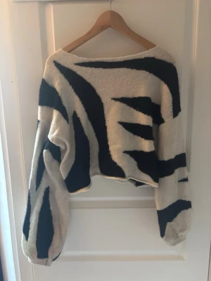 Zebramönstrad stickad tröja - Säljer en croppad stickad tröja med zebramönster i svart och vit. Tröjan har bred halsringning och långa, pösiga ärmar. Materialet är mjukt och fluffigt, perfekt för dig som vill sticka ut med en trendig look.