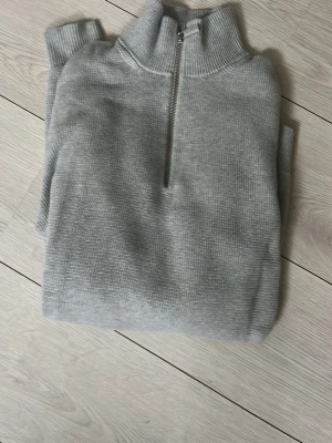 Ljusgrå stickad tröja Selected Homme - Ljusgrå stickad tröja från Selected Homme med halv dragkedja och hög krage. Tröjan har en mjuk struktur och ribbstickade detaljer vid halsen. Perfekt för lager på lager och enkel att matcha med olika outfits.
