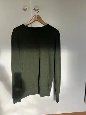 Mörkgrön stickad tröja - En enkel och stilren mörkgrön stickad tröja med rund halsringning och långa ärmar. Tröjan har en diskret liten logga vid nederkanten och är tillverkad i ett mjukt, tunt material som passar perfekt till lager på lager.