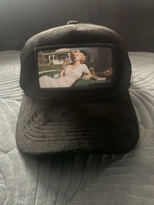 Svart truckerkeps Maggiore - Säljer en svart truckerkeps från Maggiore med mesh på sidorna och ett stort färgfoto framtill som motiv. Kepsen har böjd skärm och justerbar baksida. Perfekt för dig som gillar streetwear och unika accessoarer.