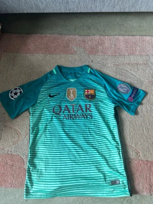 FC Barcelona fotbollströja - Säljer en turkos FC Barcelona matchtröja från Nike med Qatar Airways-tryck, klubbmärke och Champions League-patch på ärmen. Tröjan har korta ärmar, rund hals och tunna horisontella ränder framtill. Perfekt för fotbollsträning eller att bära som supporter.
