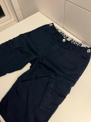 Mörkblå cargo shorts från BOSS - Snygga mörkblå cargo shorts från BOSS med flera praktiska fickor på sidorna och bak. Shortsen har en tydlig BOSS-logga på insidan av midjan och är tillverkade i ett mjukt bomullsmaterial. Perfekta för en avslappnad och trendig stil.